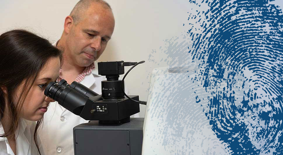 Image: Teacher in a White Coat Overseeing a Female Student in a White Labcoat peering into a microscope