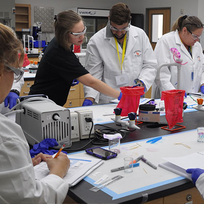 Image: Four people in white coats and lab gear working at a table with red biohazard bags, scanning equipment, and vials. One person is filling out a worksheet in pencil.