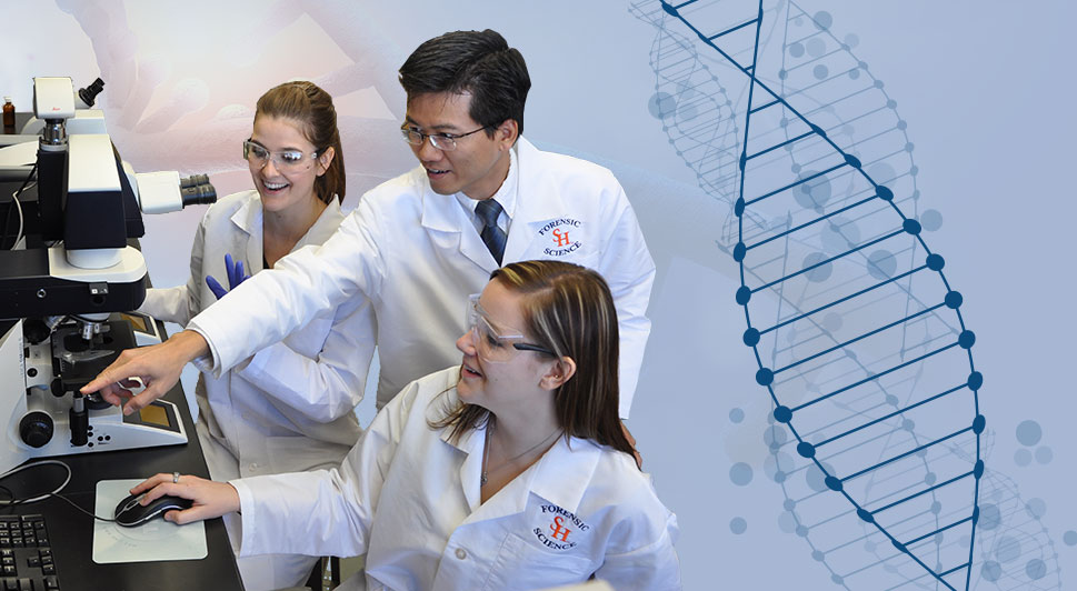 Image: Three individuals in white lab coats seated by a large microscope and computer screen. The middle individual is pointing offscreen towards the computer.